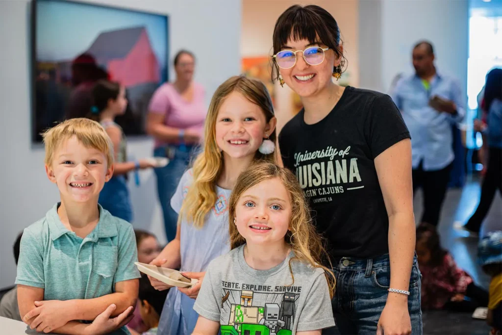 A smiling adult in a University of Louisiana shirt and three children pose indoors at the Hilliard Art Museum, surrounded by others. The group is beaming, highlighting the joy found in Louisiana museums and local art Lafayette offers.