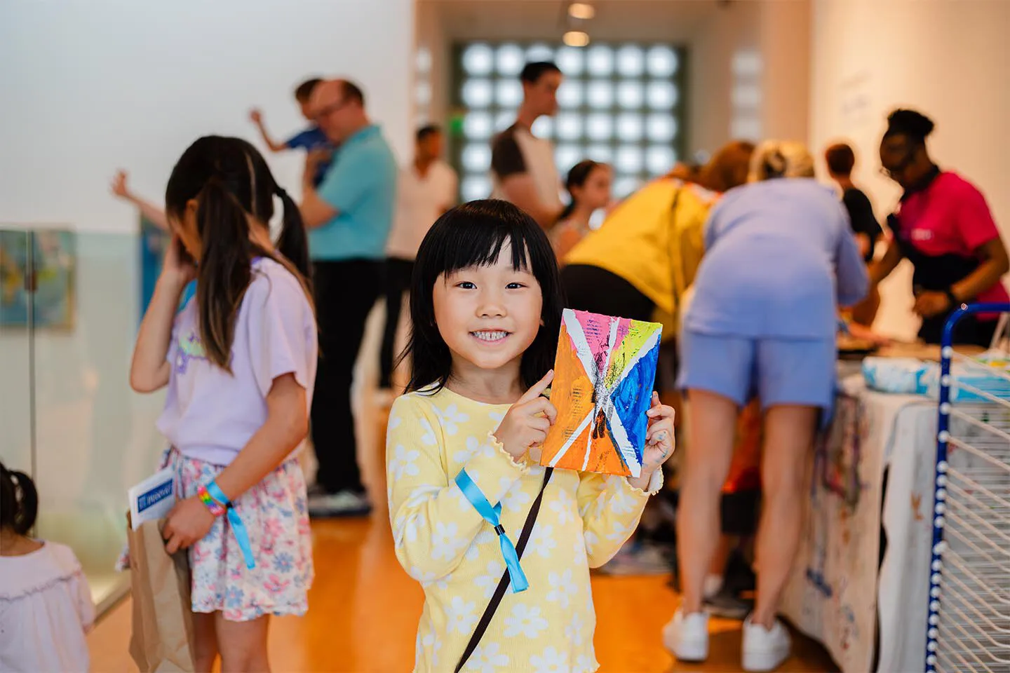 A young girl smiles and holds up a colorful piece of artwork. Behind her, children and adults participate in an art activity at the brightly lit Hilliard Art Museum, a hub for local art in Lafayette.
