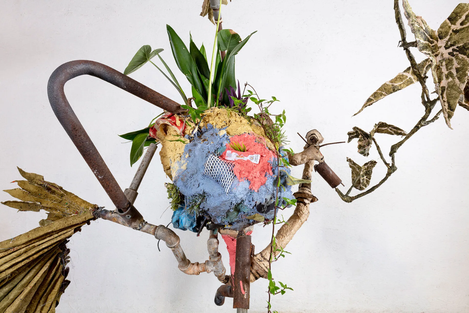 Mixed-media sculpture with a heart-shaped form of colorful fabric, netting, and live plants attached to metal pipes and branches—all set against a plain white background—perfect for display at a Lafayette art museum or Louisiana museums.