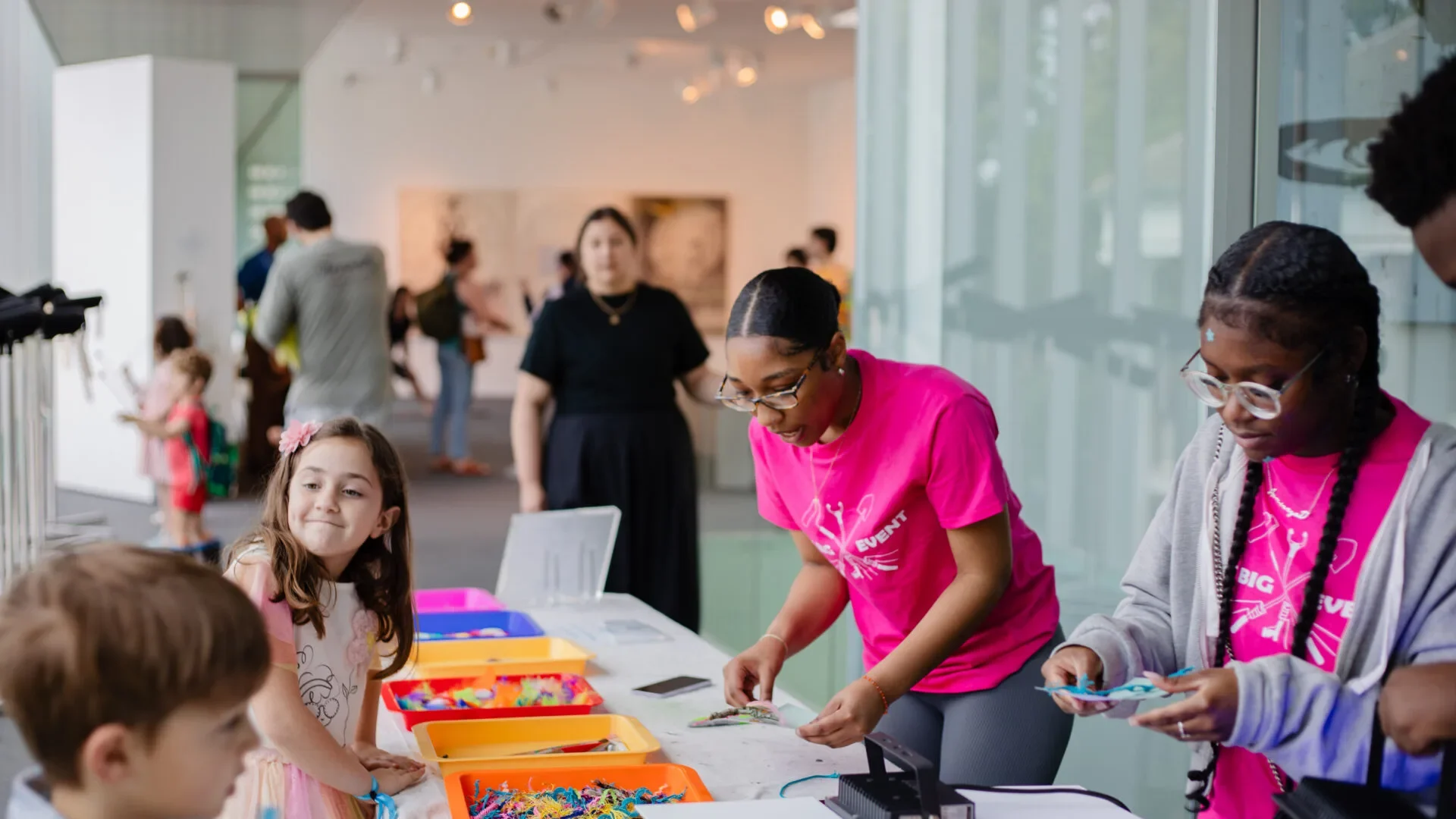 A group of children and adults gather around a table filled with colorful craft supplies, making crafts together in a bright, modern indoor space at the Hilliard Art Museum. One adult in a pink shirt helps guide the activities during Hilliard exhibitions.