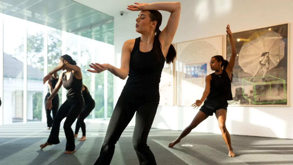 Four women in black dance outfits perform contemporary dance moves in a bright, modern room at the Lafayette art museum, with large windows and framed artwork on the walls. The dancers are mid-motion, expressing emotion through their poses.
