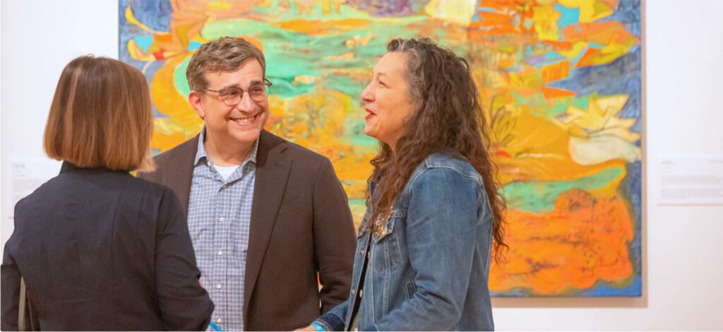 Three people stand and chat, smiling in front of a colorful, abstract painting at the Lafayette art museum. Two face each other while one has their back to the camera, creating a casual and friendly scene amid vibrant local art Lafayette displays.