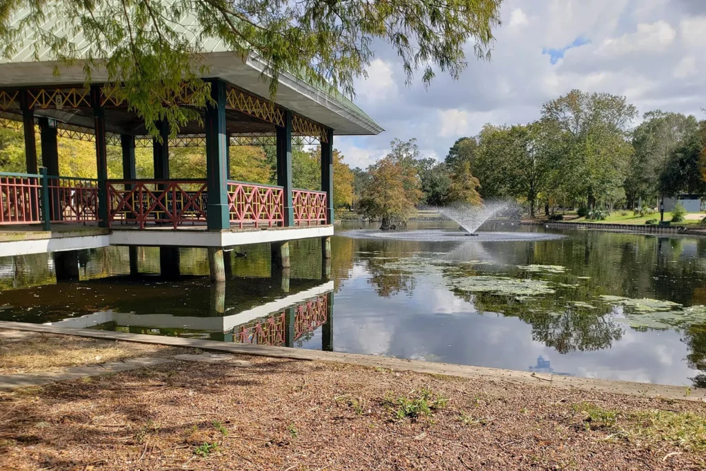A pavilion with red railings extends over a calm pond and central water fountain, surrounded by green trees under a partly cloudy sky at the family friendly Hilliard Art Museum, home to engaging Hilliard exhibitions in Lafayette.