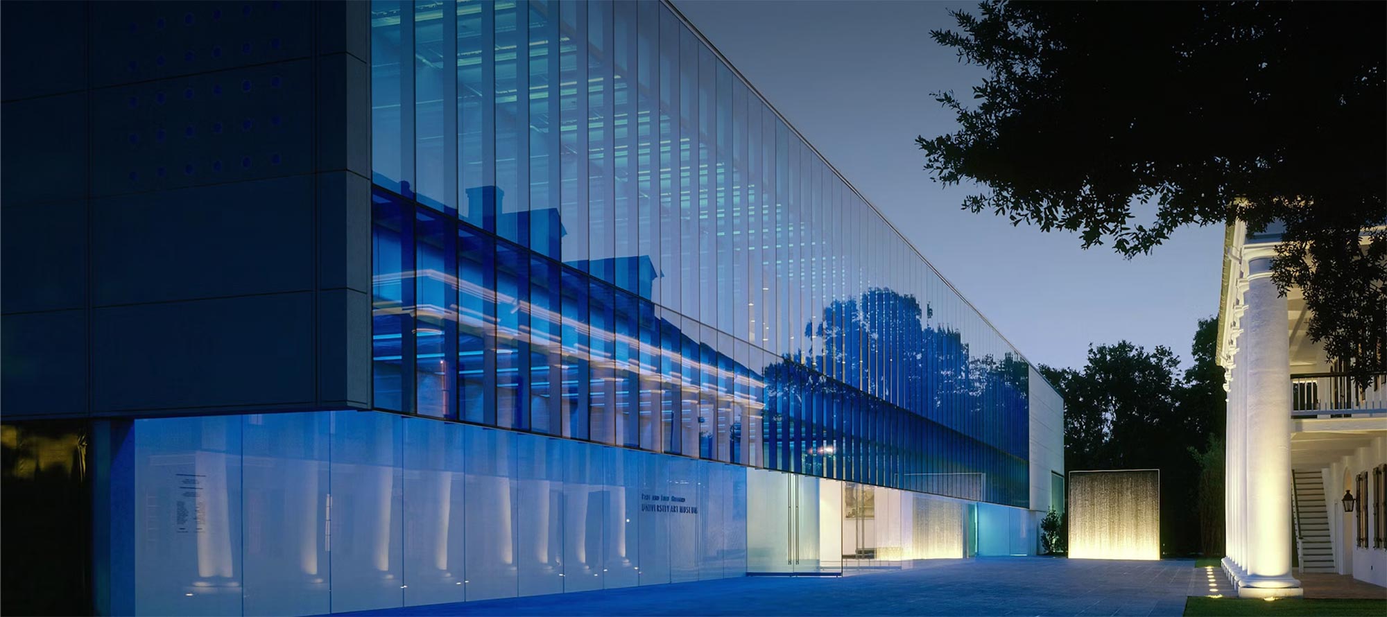 A modern glass building glows with blue light at dusk, reflecting trees and another classical-style building with columns on the right. This serene scene captures the elegance of Louisiana museums and local art Lafayette.