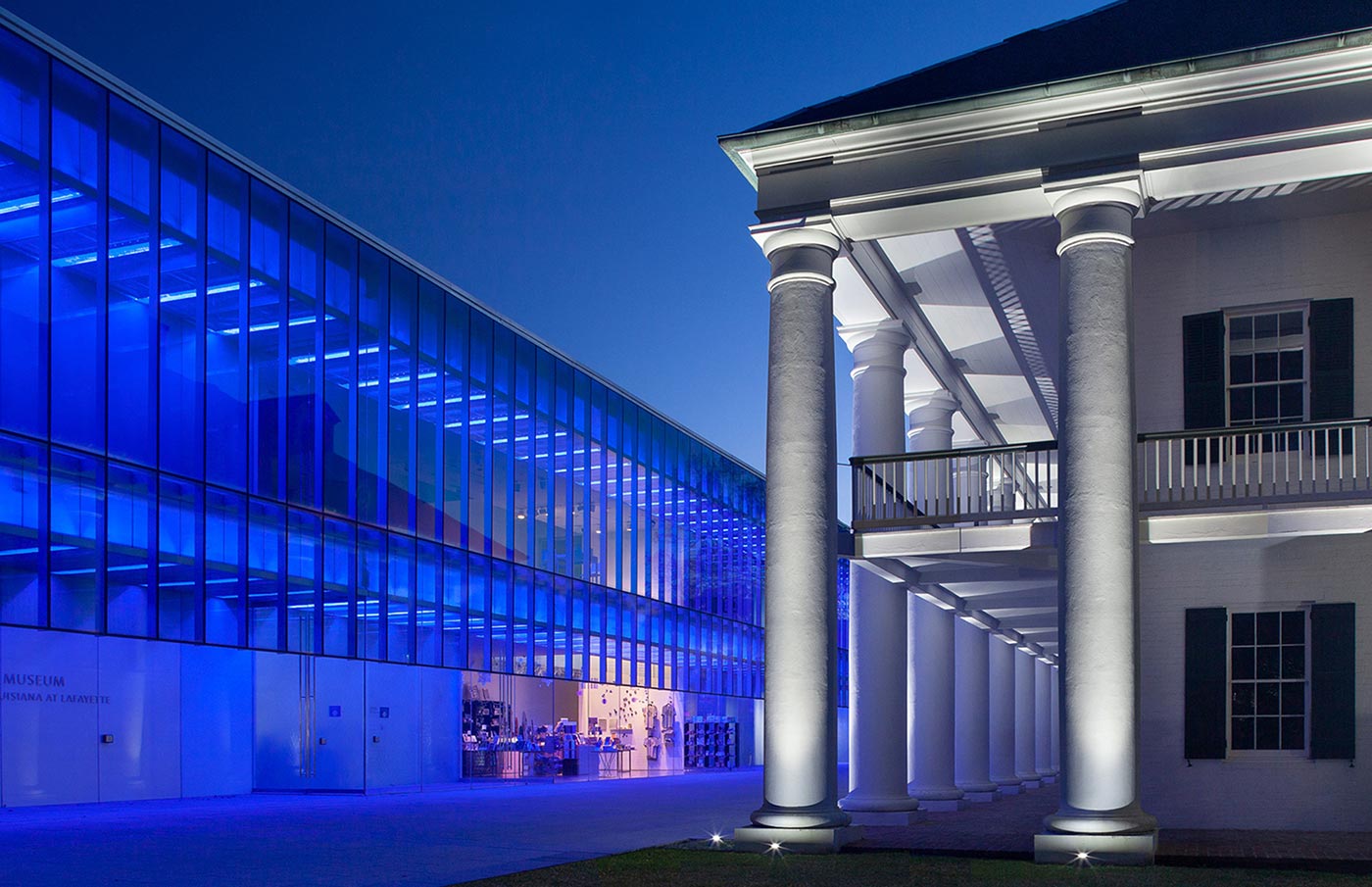 A modern glass building lit with blue lights stands beside a classic white columned building at dusk, showcasing a striking contrast in architectural styles—an inviting scene typical of Louisiana museums and Lafayette family friendly attractions.
