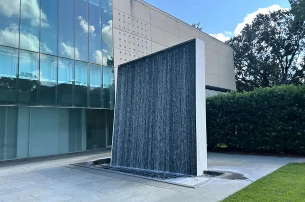 A modern outdoor water fountain features a tall, rectangular wall with water cascading down its surface, beside a glass building at the Hilliard Art Museum and lush green hedges under a blue sky—perfect for enjoying local art Lafayette exhibitions.