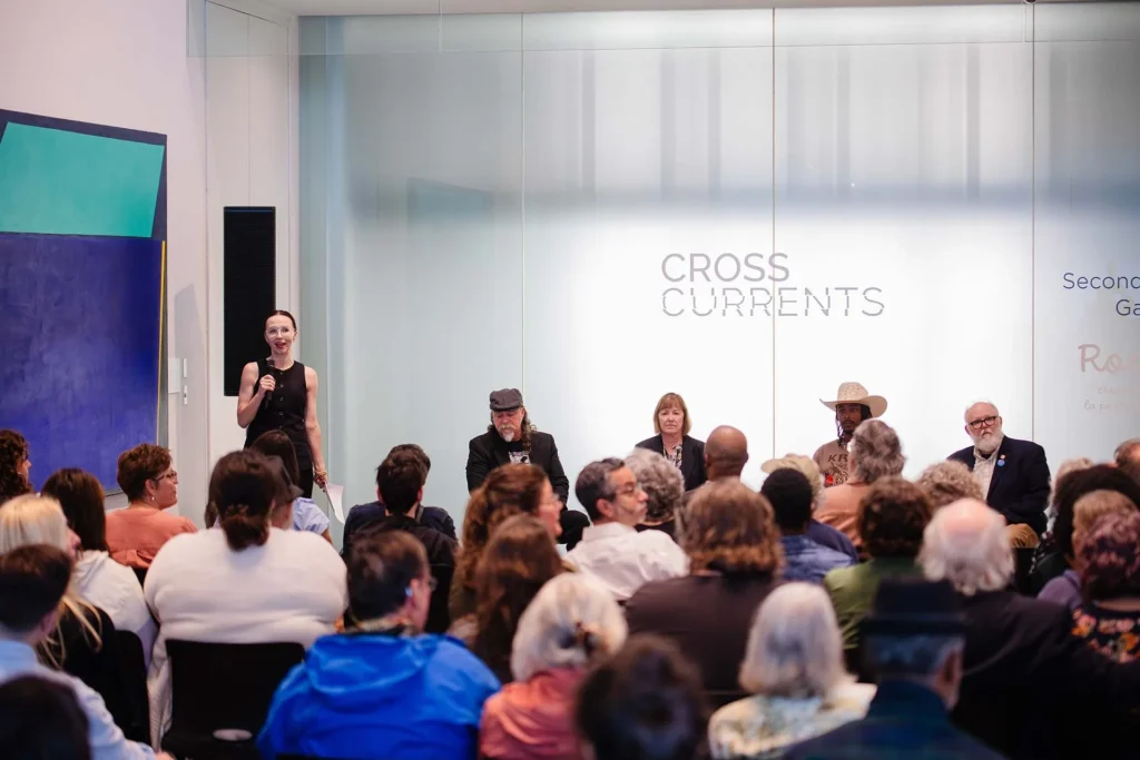 A woman stands and speaks to an audience at the Hilliard Art Museum. Four people sit on a panel beside her, with a CROSS CURRENTS sign behind them. The room, filled with attendees, reflects the Lafayette family friendly spirit of Louisiana museums.