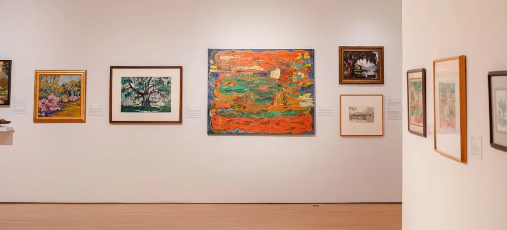 A gallery wall displays various framed artworks, including colorful paintings and sketches, arranged in a row on a white wall with wooden flooring visible beneath.