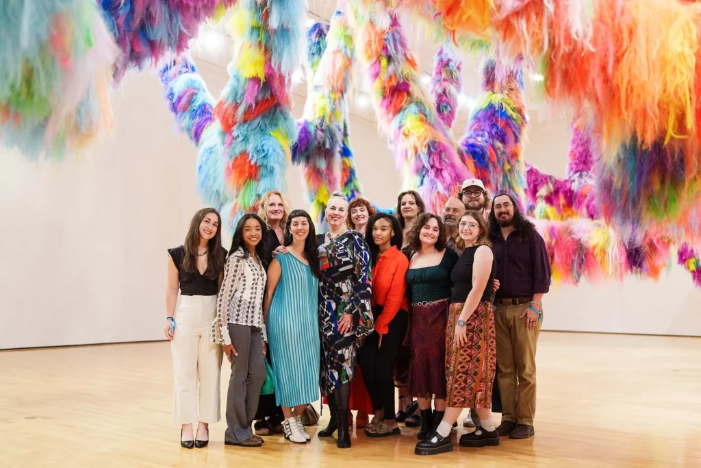 A group of people stands smiling together in a gallery, surrounded by colorful, hanging, fluffy art installations in a bright, spacious room with wooden floors.
