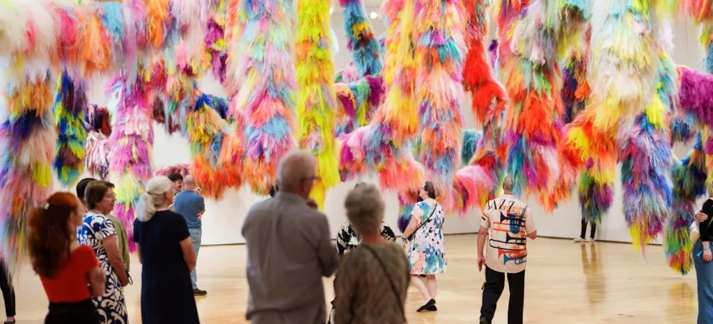 A group of people observes a colorful art installation featuring large, hanging clusters of vibrant, fluffy fibers in various bright colors inside a spacious gallery.