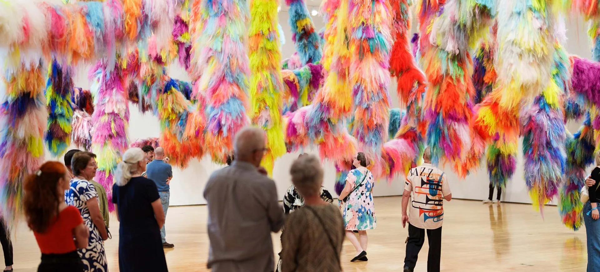 A group of people observes a colorful art installation featuring large, hanging clusters of vibrant, fluffy fibers in various bright colors inside a spacious gallery.