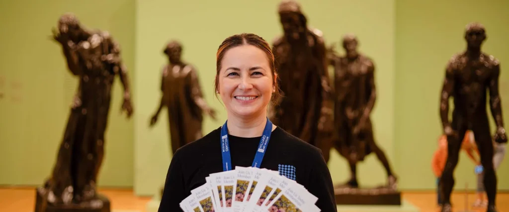 A smiling woman with a lanyard holds several brochures, standing before large dark bronze statues in the Hilliard Art Museum—a Lafayette family friendly destination with a light green gallery wall.