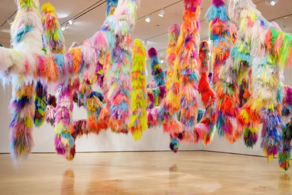 Colorful, fluffy, fur-like sculptures hang from the ceiling in a gallery space with wooden floors and white walls, creating a vibrant and playful art installation.