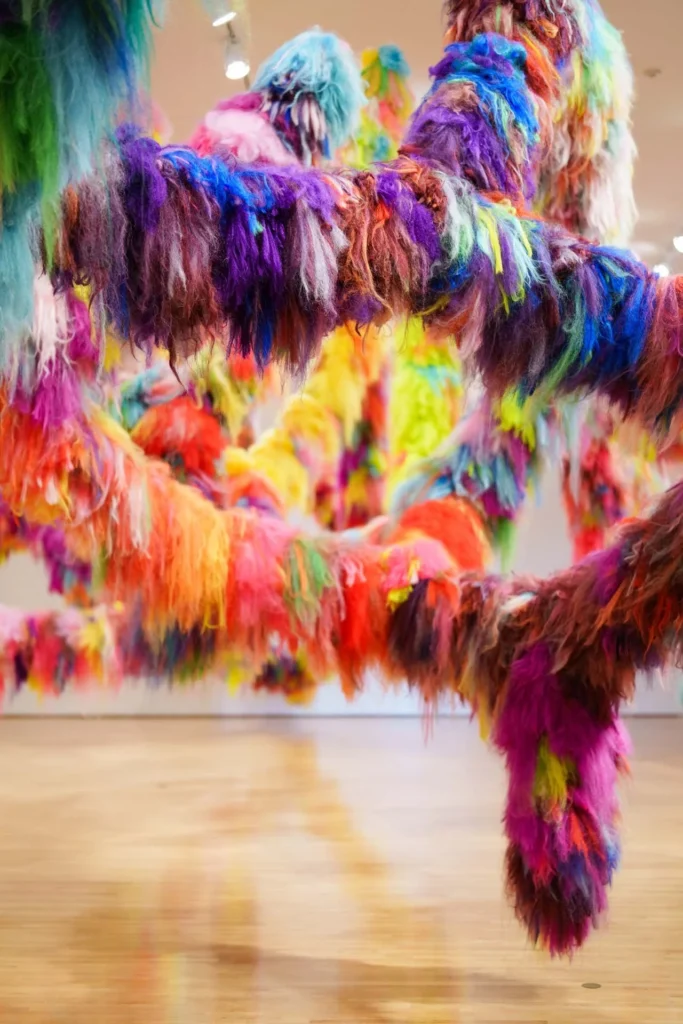 Bright, colorful, furry strands in shades of pink, purple, orange, green, blue, and yellow hang from the ceiling in an art gallery, creating a whimsical, immersive installation over a polished wooden floor.