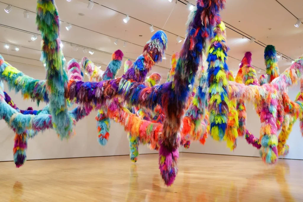 Colorful, fluffy, tentacle-like sculptures hang from the ceiling in an art gallery, filling the space with vibrant hues of blue, pink, yellow, green, and orange against white walls and a shiny wood floor.