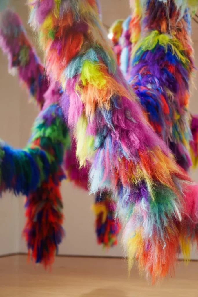 Colorful, furry, branch-like sculptures hang in an art gallery space, featuring long, shaggy fibers in vibrant rainbow hues, including pink, blue, green, yellow, orange, and purple.