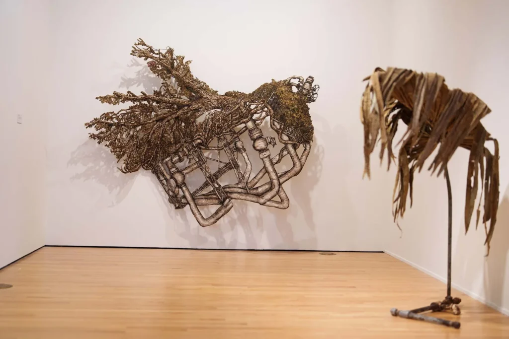 A metal and organic branch-like sculpture hangs on a white wall, with a second sculpture of twisted, leaf-like materials standing on the wooden gallery floor to the right.