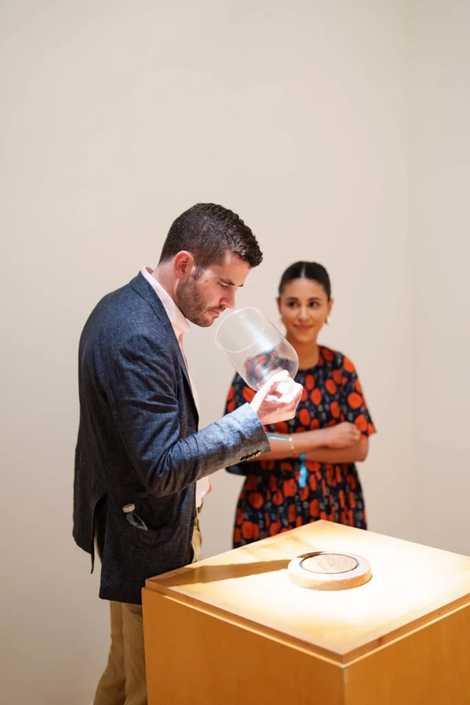 A man in a suit jacket smells inside a large glass vessel, while a woman in a patterned dress stands behind him, smiling, near a wooden display pedestal in a brightly lit room.