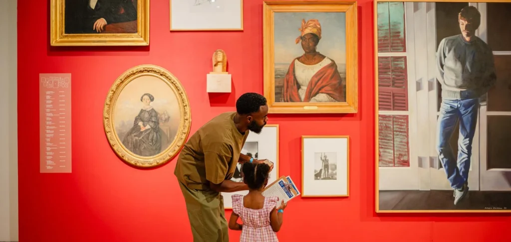 A man and a young girl admire paintings at the Hilliard Art Museum, surrounded by portraits in gold and white frames on a bright red wall. The girl holds a pamphlet while the man gestures toward artworks from Hilliard exhibitions.