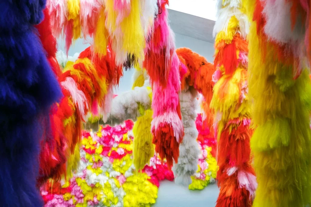 Long, colorful, fluffy fabric strips in pink, red, yellow, orange, and white hang from above at the Hilliard Art Museum, with more fluffy textures on the floor creating a vibrant, immersive, and playful environment unique among Louisiana museums.