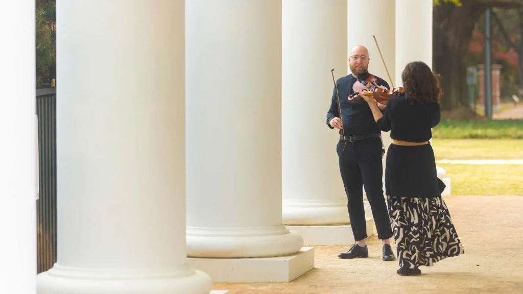 Two musicians stand outside near large white columns at the Lafayette art museum; one plays the violin while the other, holding a bow, listens. Dressed in black, they create a bright, engaging scene—perfect for a Lafayette family friendly outing.
