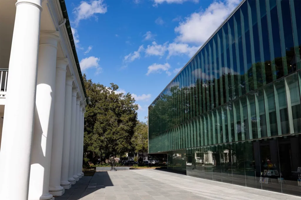 A modern glass building stands next to a traditional white columned structure under a blue sky with scattered clouds. Trees are visible in the background along the walkway between the buildings.