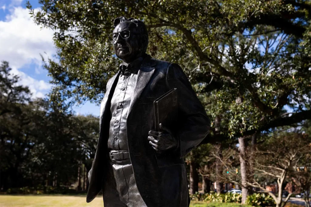 A bronze statue of a man wearing glasses and a bow tie, holding a book under his arm, stands outdoors in front of leafy trees and greenery on a sunny day.