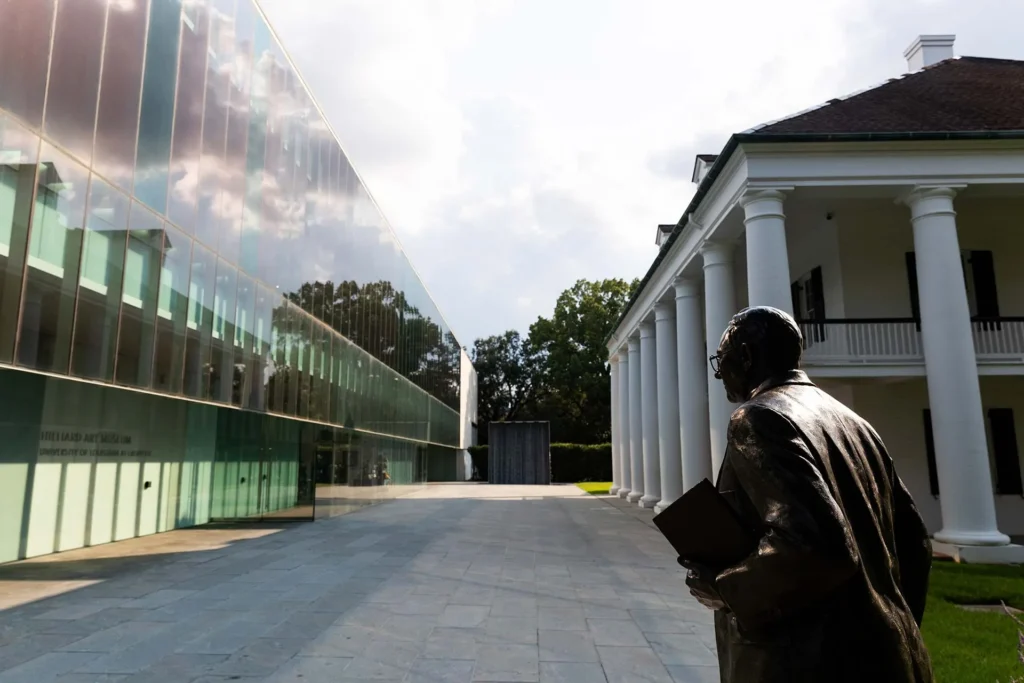 A bronze statue of a man holding a book stands in front of a historic white-columned building beside a modern glass museum; trees and sky are visible in the background.