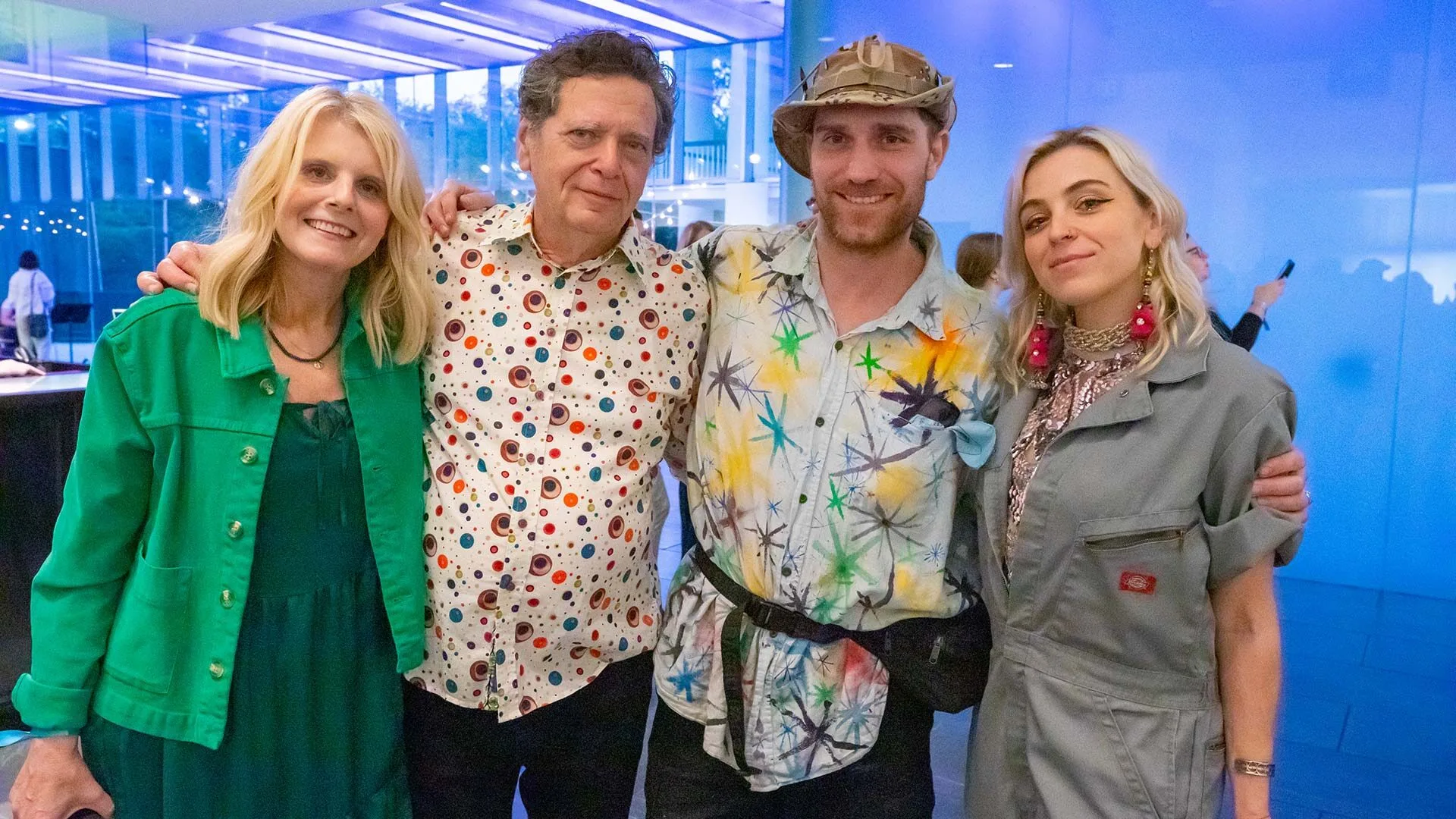 Four adults stand together indoors, smiling at the camera. Each is dressed in colorful, festive attire, perfect for the vibrant local art Lafayette scene. The modern blue-toned glass walls suggest they might be attending Hilliard exhibitions nearby.