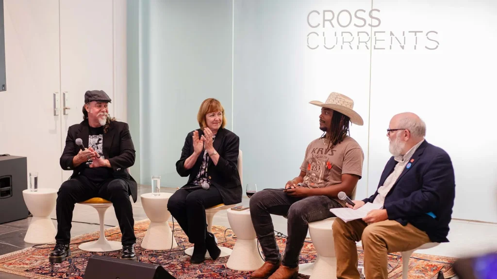 Four people sit on stools in front of a CROSS CURRENTS sign, engaged in a lively panel discussion at the Hilliard Art Museum. Two men hold microphones, a woman claps, and another man wears a cowboy hat. A rug and glasses of water are visible.