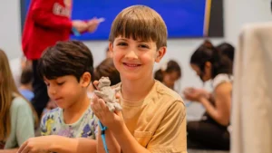 A smiling boy holds up a small clay sculpture in a classroom setting at one of Lafayette's family friendly Louisiana museums, surrounded by other children engaged in similar creative activities.