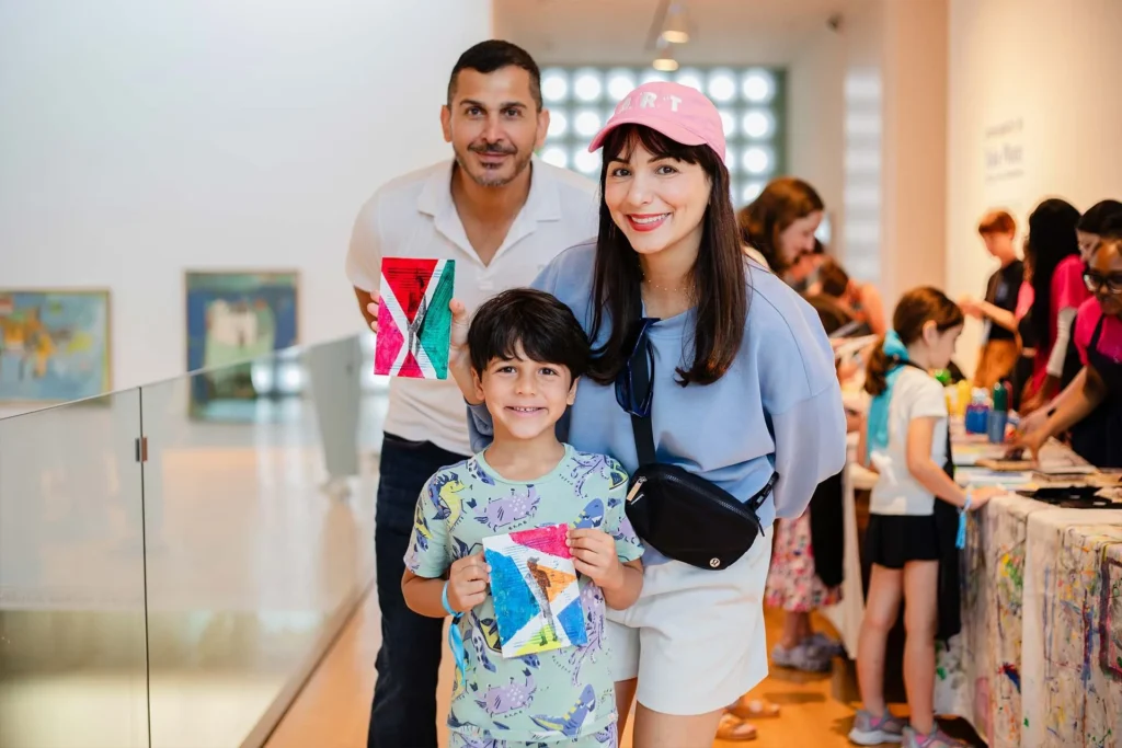 A smiling family of three poses indoors at a Lafayette art museum, holding colorful geometric art. The mother wears a pink hat, and their young son grins in front. In the background, people gather at tables, enjoying this Lafayette family friendly event.