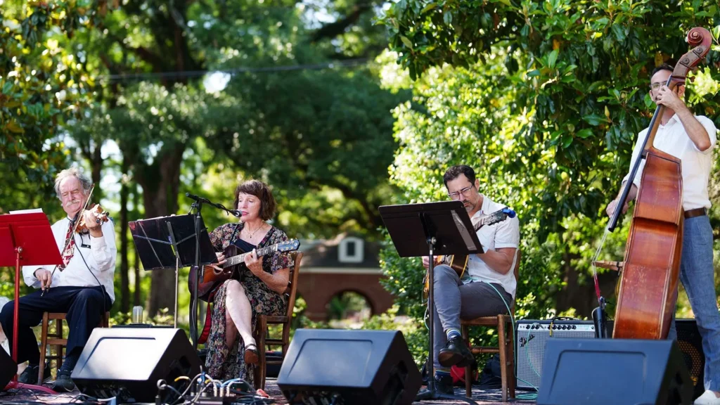 Four musicians perform outdoors on a stage surrounded by trees at a Lafayette family-friendly event. One plays violin, one sings and plays guitar, another plays clarinet, and one plays upright bass. Music stands and audio equipment are visible.