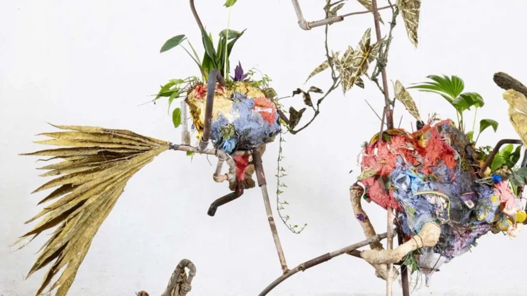 A sculptural arrangement featuring branches, dried leaves, colorful paper mache forms, and green plants—blending organic and artificial elements—evokes the innovative spirit found at Hilliard Art Museum in Lafayette. Set against a plain white background.