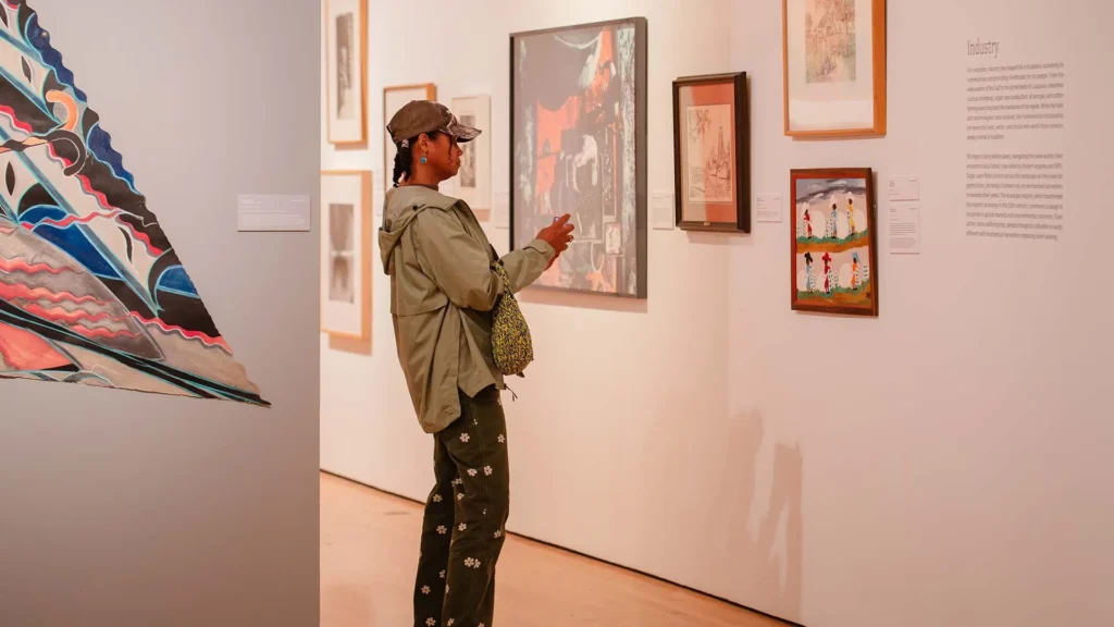 A person wearing a cap, green jacket, and patterned pants stands in the well-lit Hilliard Art Museum, looking at framed artworks on the wall and holding a phone. The gallery showcases a variety of local art Lafayette is known for.