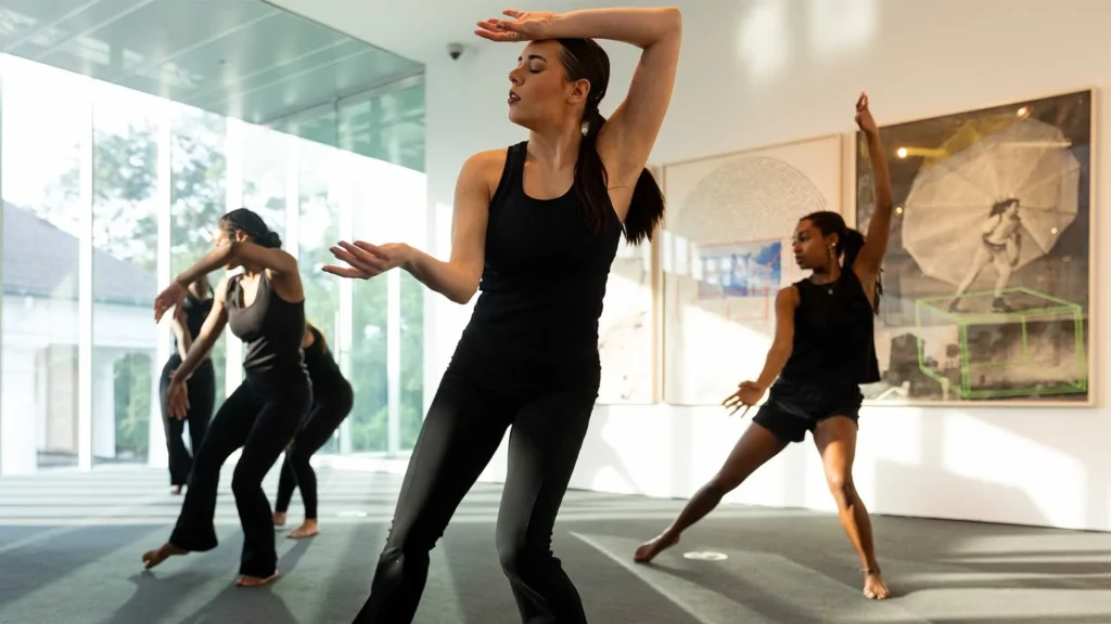 Four women in black dance outfits perform expressive contemporary dance moves in a bright, modern gallery space with large windows and framed artwork on the walls.