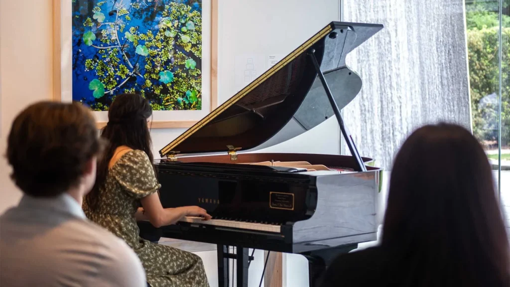 A woman in a green dress plays a grand piano indoors, while two people sit and watch. A colorful abstract painting hangs on the wall, and large windows reveal a view of trees and a waterfall outside.