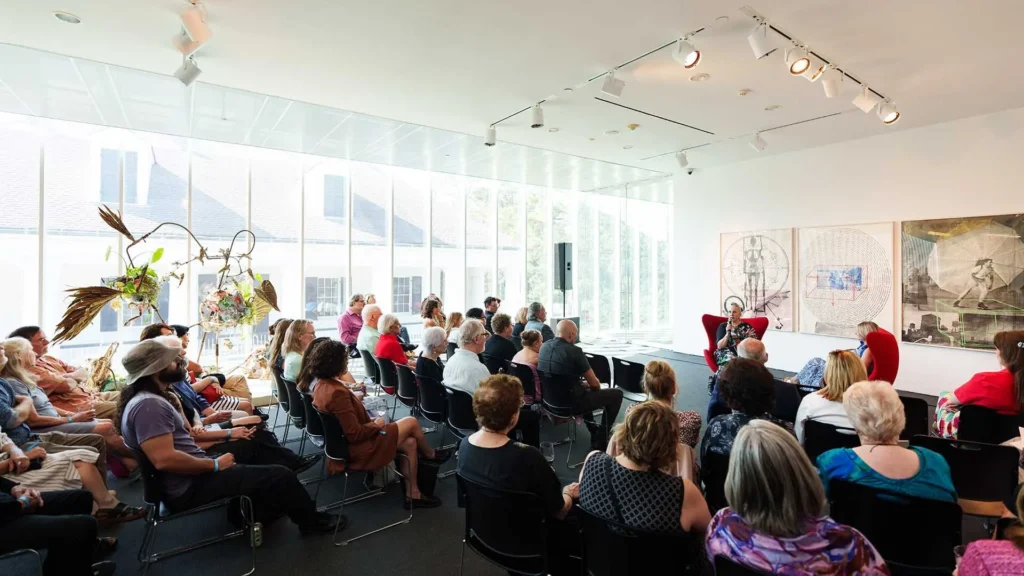 A group of people seated in a bright, modern gallery room listen to a speaker at the front. Large windows let in natural light, and artwork is displayed on the walls.