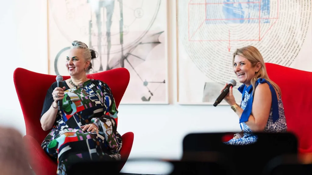 Two women sit in red chairs, smiling and speaking into microphones during an event. Abstract art is displayed on the wall behind them. One woman wears a colorful dress, the other wears a blue and white outfit.