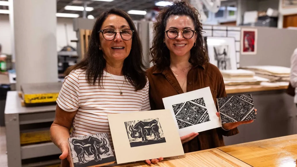 Two women indoors smile at the camera while holding up black-and-white printmaking artworks featuring intricate designs, including an elephant. Art supplies and prints are visible on the table and in the background.