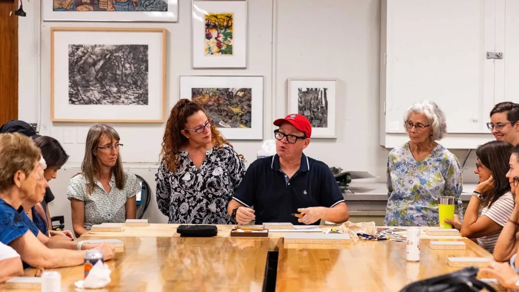 A group of adults sits and stands around a large table in an art studio, listening to a man in a red cap who is speaking and gesturing with a pen. Artworks hang on the white walls behind them.