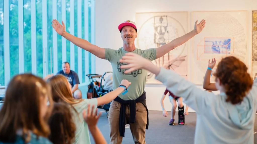 A man with outstretched arms enthusiastically leads a group of children in an indoor activity at an art gallery, with colorful artwork and large windows in the background.