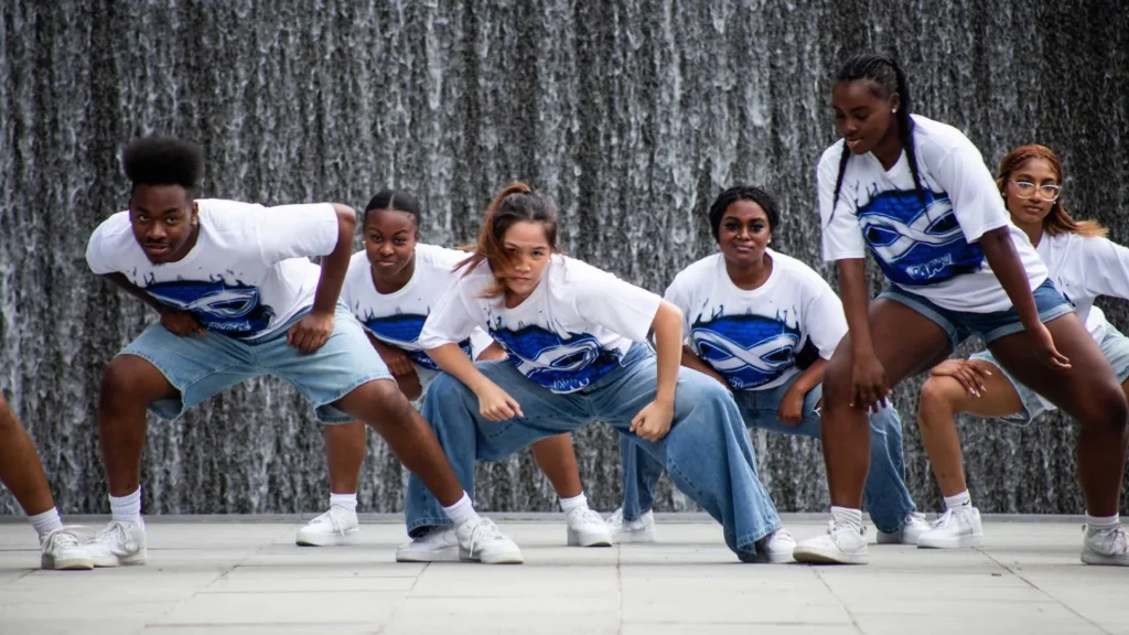 Five people wearing matching white and blue shirts and denim pose in a low, energetic dance stance in front of a large, cascading waterfall.