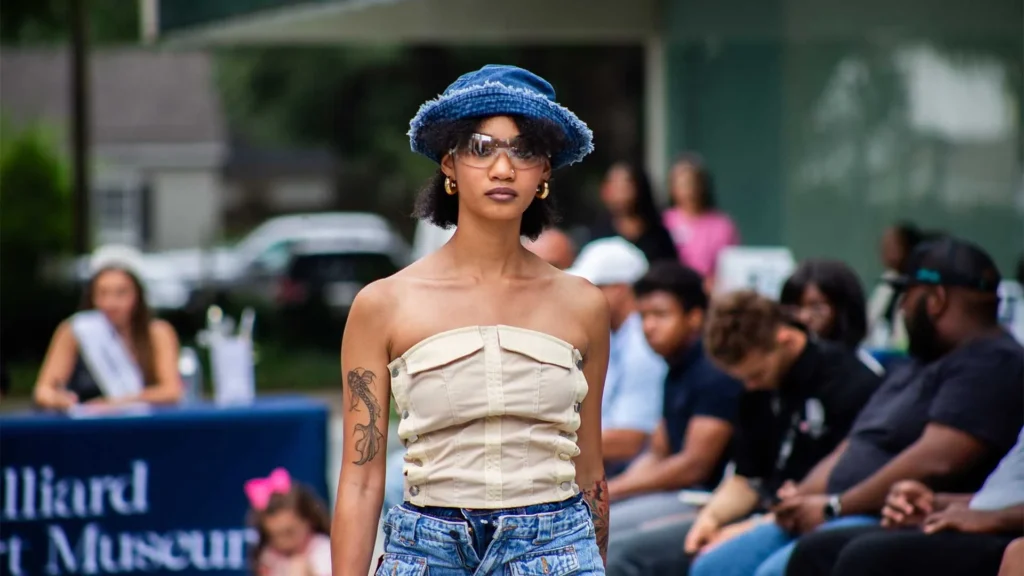 A model walks outdoors wearing a strapless beige top, blue denim bucket hat, sunglasses, and patterned jeans. People are seated and watching in the background, with a sign for an art museum visible.
