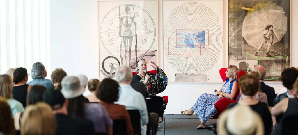 A seated audience listens to two people engaged in conversation on stage in front of large abstract artworks, with one person gesturing while speaking and the other seated in a blue dress.