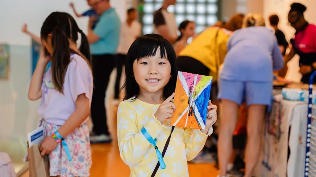 A young girl in a yellow dress smiles and holds up a colorful piece of art at the Hilliard Art Museum, with other children and adults enjoying the lively atmosphere in the background.