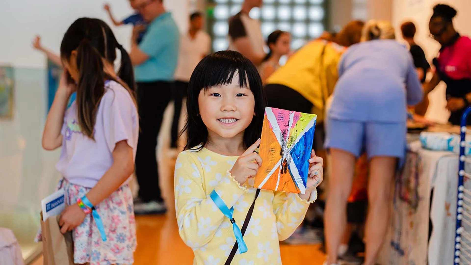 A young girl in a yellow dress smiles and holds up a colorful piece of art at the Hilliard Art Museum, with other children and adults enjoying the lively atmosphere in the background.