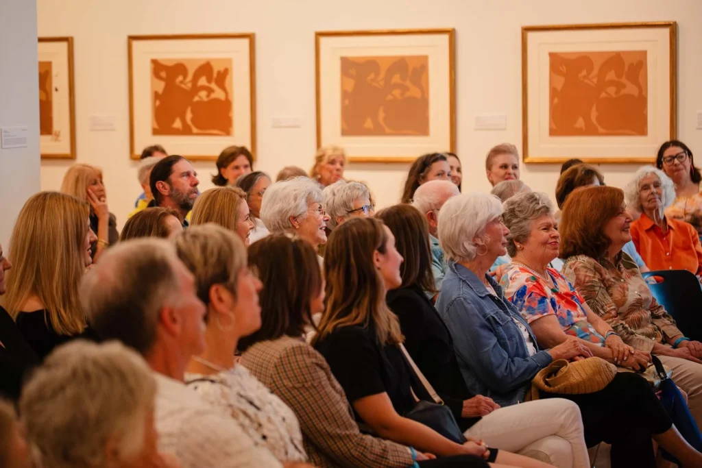A group of people of various ages sit closely together, attentively watching an event at the Lafayette art museum, where abstract artwork lines the walls and the atmosphere feels inviting and Lafayette family friendly.