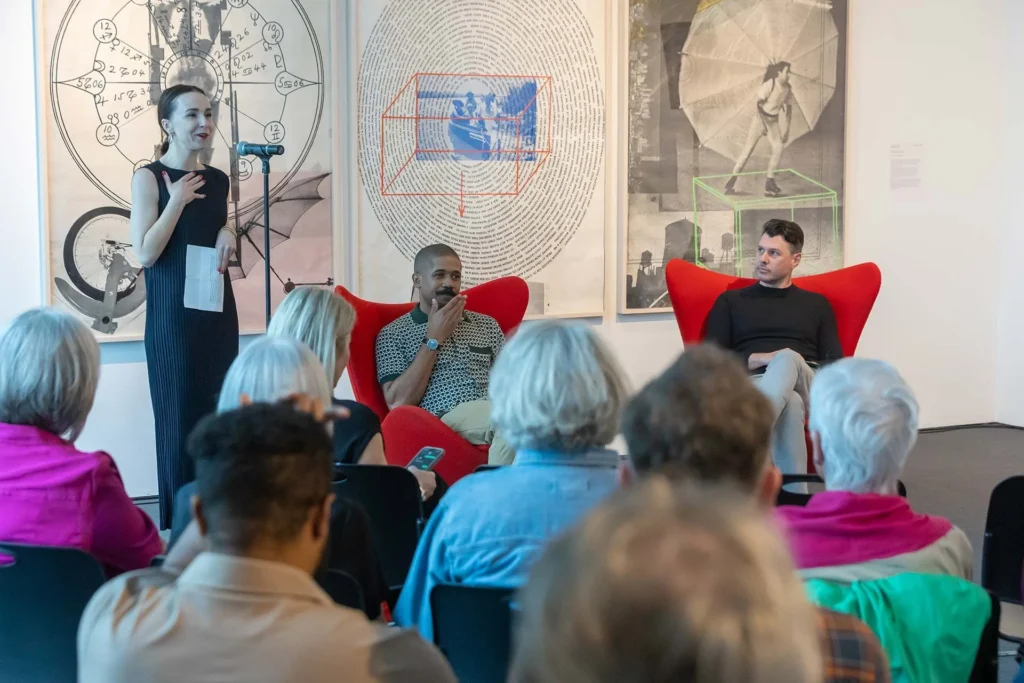 A woman speaks at a microphone to an audience in an art gallery, while two men sit in red chairs behind her. Large contemporary artworks, showcasing local art Lafayette, hang on the wall in the background.