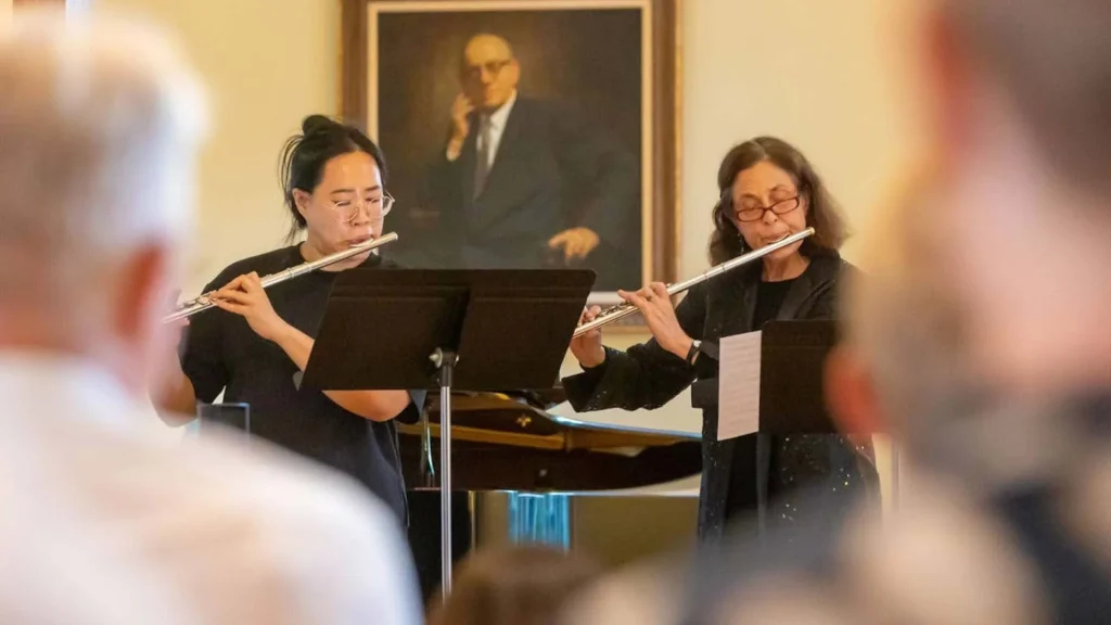 Two women play flutes side by side in front of music stands, with a piano behind them and a blurred audience in the foreground. A framed portrait of a seated man hangs on the wall in the background.